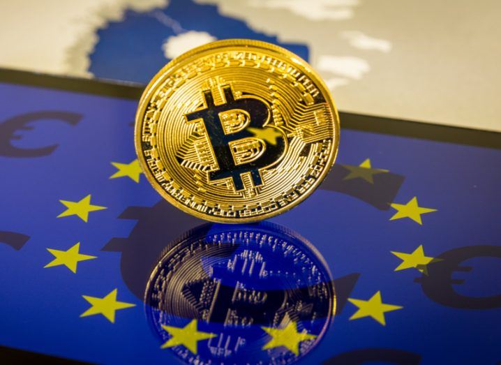 A gold coin with the bitcoin logo, on top of a mobile phone that has the EU flag and euro symbols on its screen. The phone is on top of a map of Europe.