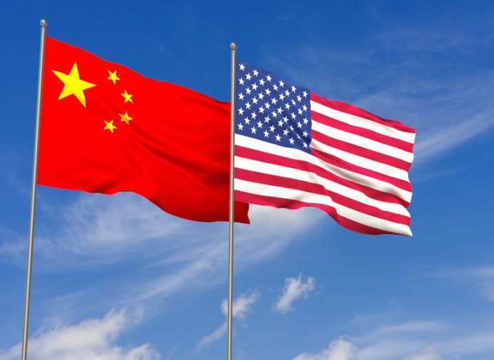 The flags of the US and China on flag poles next to each other, with a blue sky and clouds in the background.