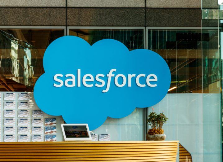 The Salesforce logo on a glass building wall, with a wooden desk in front of it. A potted plant and a small screen are on top of the desk.