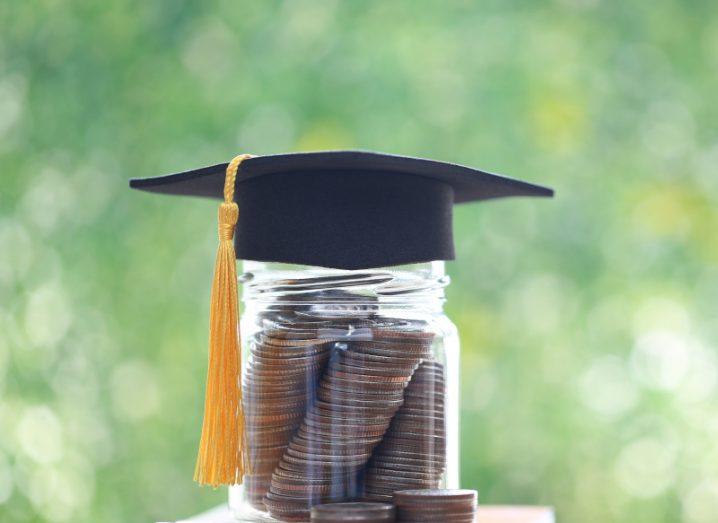 A glass jar full of coins with a graduation hat on top of it, with a blurred green background. Used to represent education funding.