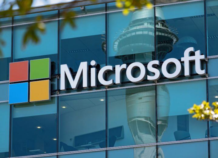 The Microsoft logo on the front of a windowed building, with another building reflected in the windows and leaves visible near the top of the image.