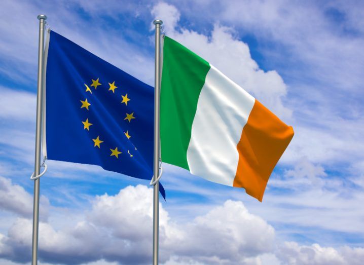 The Irish flag in front of the EU flag, both of which are on flag poles with a cloudy sky in the background.