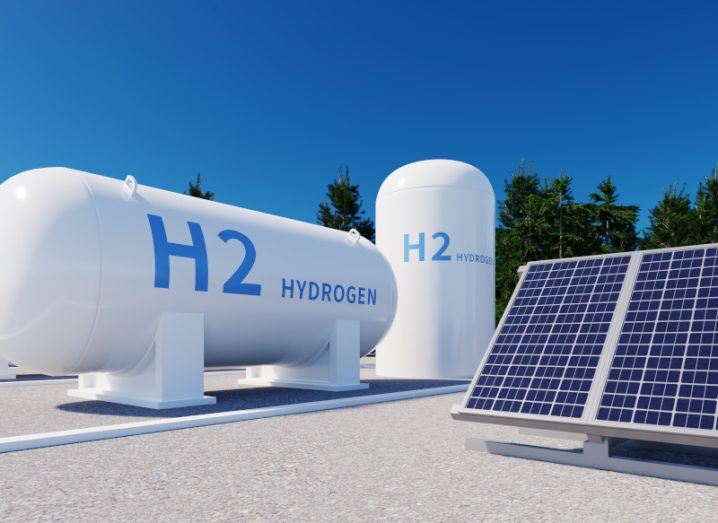 Two white storage tanks that have 'H2 Hydrogen' written on the side, next to a solar panel. There are trees and a clear blue sky in the background.