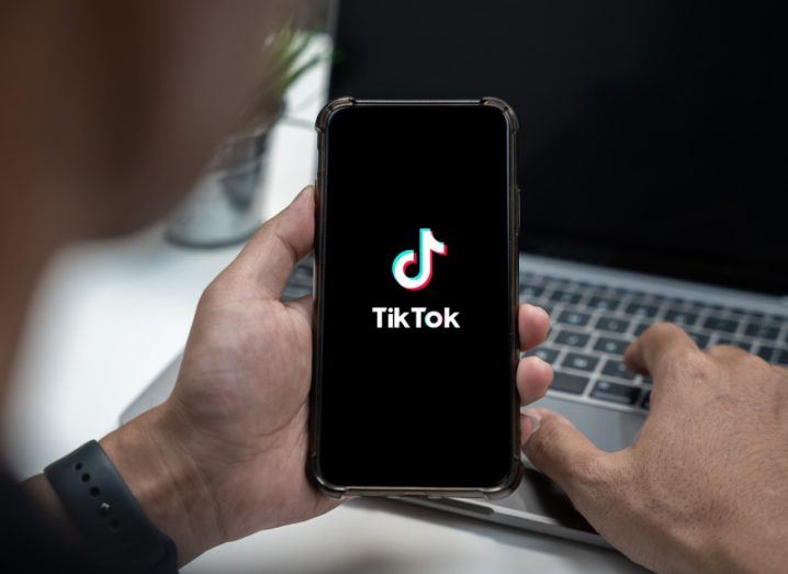 The TikTok logo on a smartphone screen, held in a person's hand in front of a laptop.