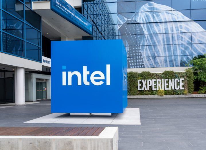 The Intel logo on a blue square outside of a building. A row of green hedges is in the background with the word "Experience" written in front of the hedges.