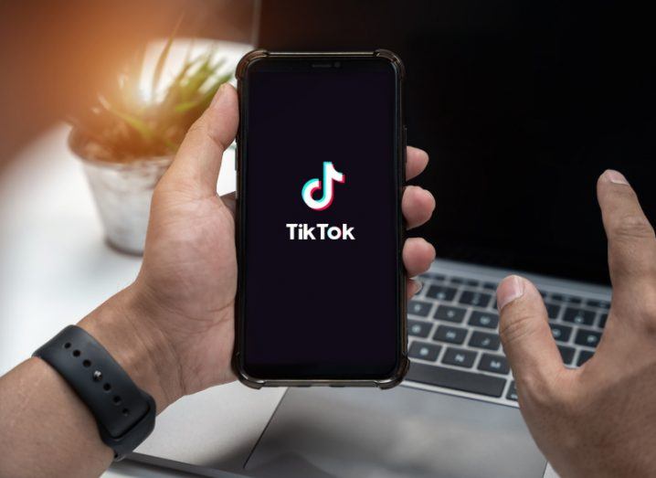The TikTok logo on a smartphone screen, which is in a person's hand. The phone is in front of a desk that has a small potted plant and a laptop on it.