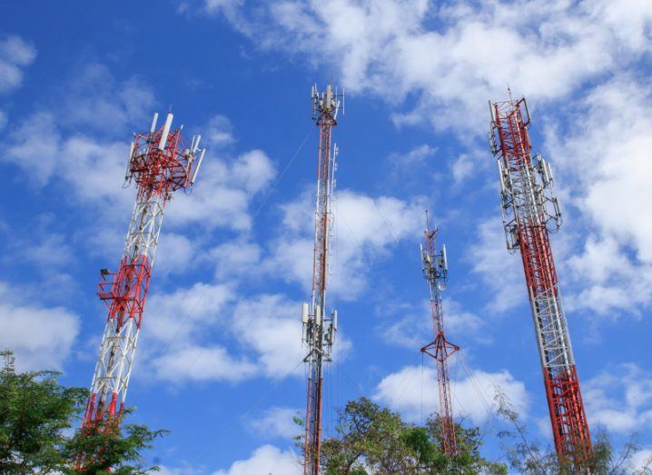 Multiple red and white communications towers, with a blue sky above them and the tops of some trees visible below.