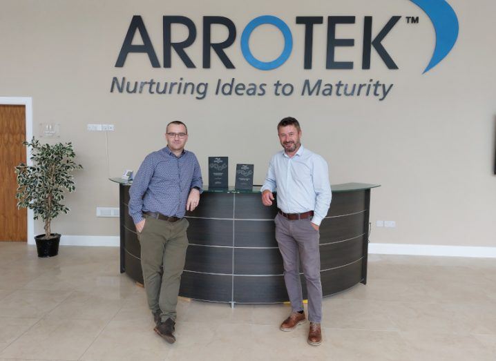 Two men standing in front of a table, with the Arrotek logo on a wall behind them. They are the co-founders of Arrotek.