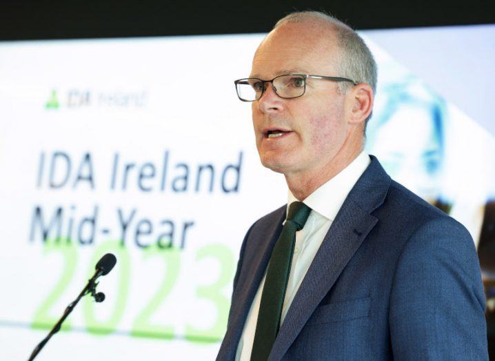 Simon Coveney, TD, in glasses and a dark suit and tie stands at a podium on a stage with IDA Ireland Mid-Year on a sign behind him.