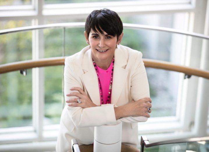 A woman wearing a white suit with a pink shirt smiles with her arms crossed while leaning on a cylindrical object. She is Carolan Lennon, country leader at Salesforce Ireland.