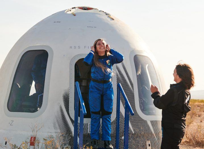 A young woman in a blue spacesuit stands outside a space craft smiling up at the sky. She is Sara Sabry.