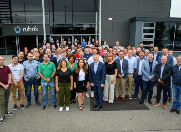A large group of people standing in front of a dark grey building that has the Rubrik logo on it.