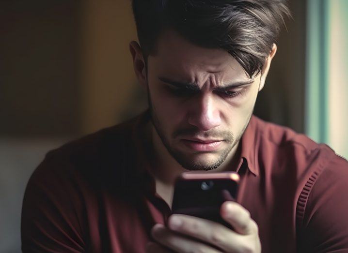Close-up of a man staring at a phone in his hand and looking distraught. Represents people using social media platforms such as Facebook and being influenced by their algorithm.