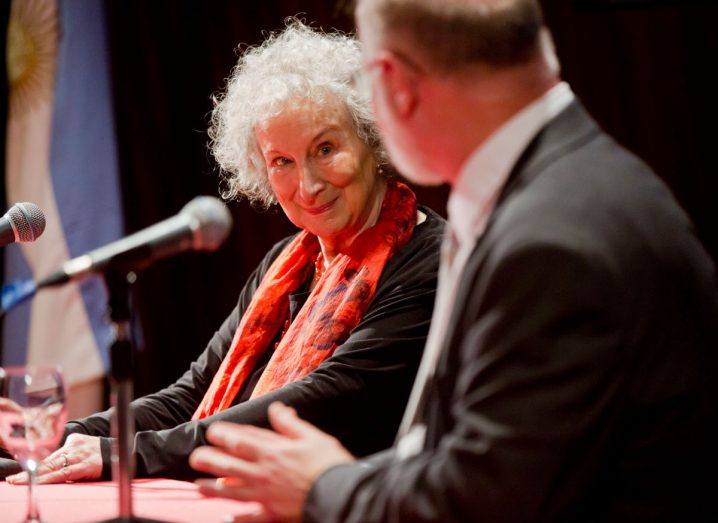 Margaret Atwood seated on stage chatting with a man with microphones in front of each of them.