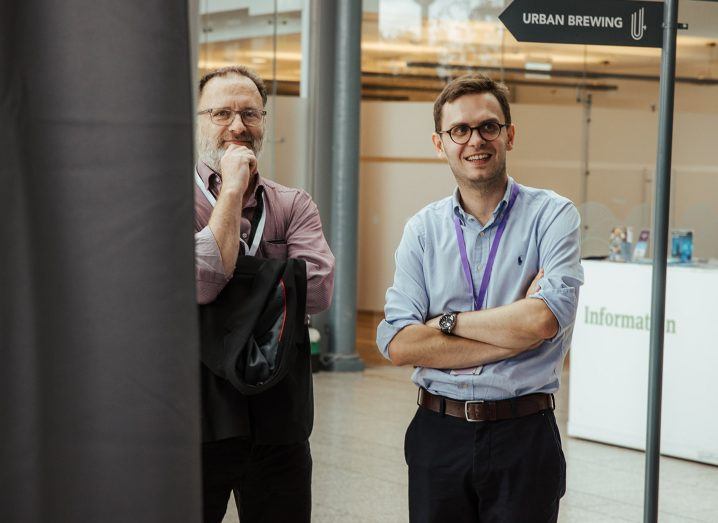 Two men standing next to each other and pose for a photo. The one on the left if partially covered by a drop down curtain at the Eyes Off Data Summit 2023 in Dublin.