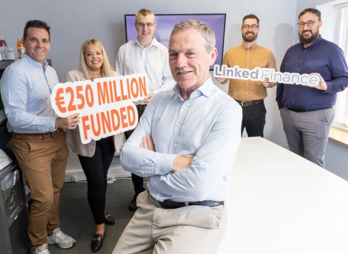 A group of people standing around a table in an office space. Man in front has arms folded and is seated on the table. Others in the background are holding cardboard cutouts that read "250 million euros" and "Linked Finance".