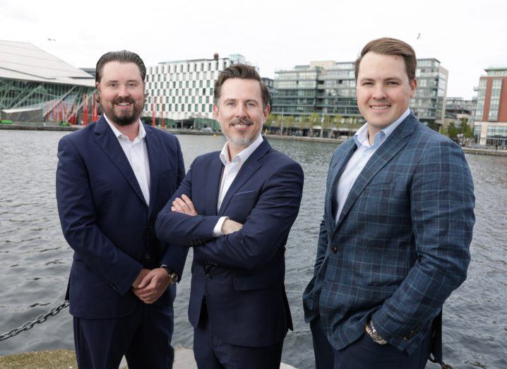 Three men wearing business formals stand in front of the Liffey in Dublin.