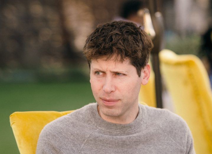 Headshot of OpenAI CEO Sam Altman wearing a grey jumper and seated in an outdoor space with yellow chairs on grass.