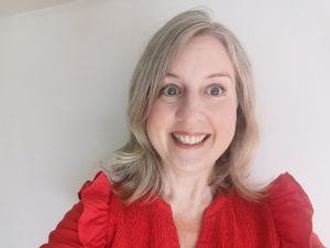 A selfie of Dr Sarah Robinson. She has blonde hair, blue eyes and is smiling and wearing a red top, standing against a cream wall.,