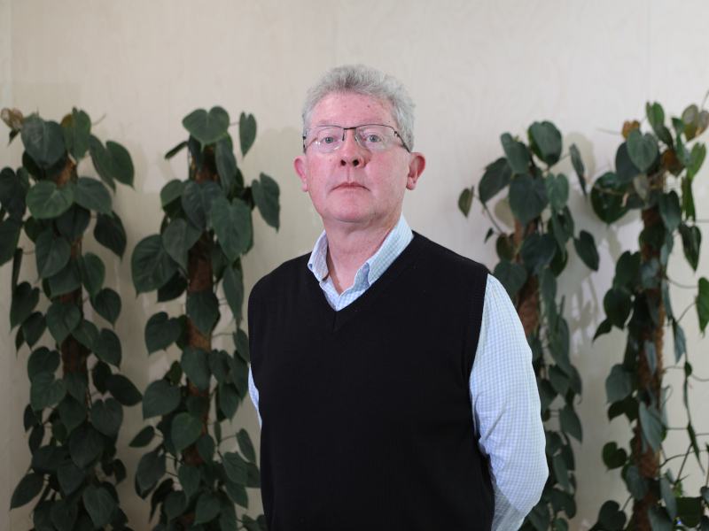 Prof Seamus Davis stands with his hands behind his back looking at the camera. He has grey hair and glasses and is wearing a blue shirt and a navy sleaveless jumper. There is a cream wall behind him with plants visible.