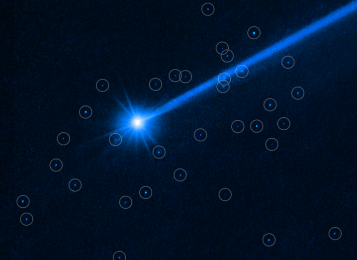 Image of a distant light with a blue tail and multiple small dots around it that have circles surrounding them. The big light is the Dimorphos asteroid.