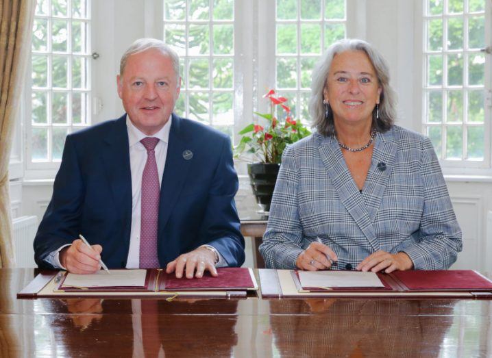 A man and a woman sit at a large boardroom table side by side. They are both holding pens and signing an agreement while smiling at the camera.