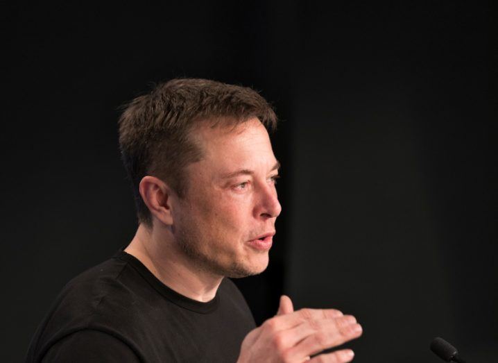Elon Musk in a black t-shirt, speaking in a dark background.