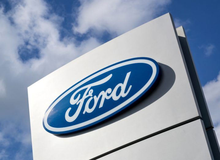 The Ford company logo on a white sign, with clouds and a blue sky above.