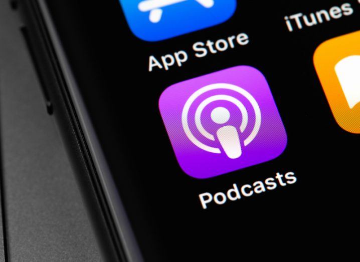 The Apple Podcasts app logo on a smartphone screen, with other apps visible next to it.
