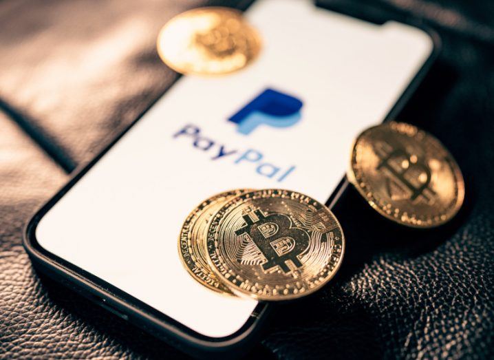 Multiple gold coins with the bitcoin logo on them, on a smartphone screen that has the PayPal logo on it.
