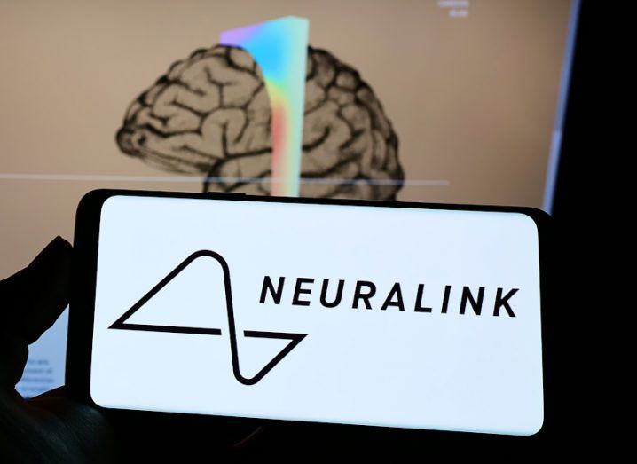 The Neuralink logo on a smartphone screen. The phone is in a person's hand, with a computer screen in the background showing an illustration of a brain.