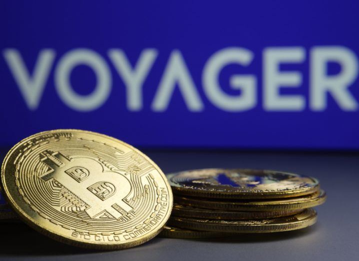 A small pile of coins with the bitcoin logo on them and the Voyager logo in the background.