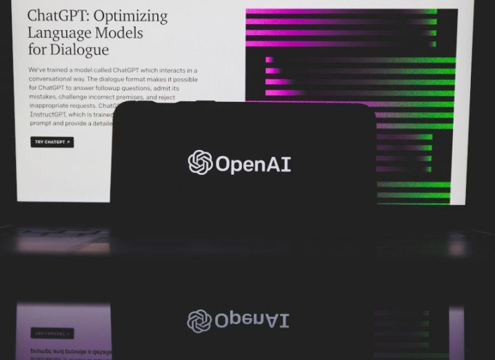 The OpenAI logo on a smartphone screen. The phone is in front of a computer screen that has the ChatGPT website on it. The phone is also on a dark, reflective surface.
