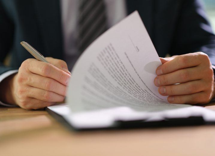A person in a suit signing a document with a pen. The document is on a wooden table.