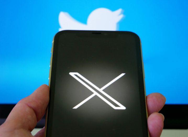 The X logo on a smartphone screen, held in a person's hand, with the Twitter logo in the background.