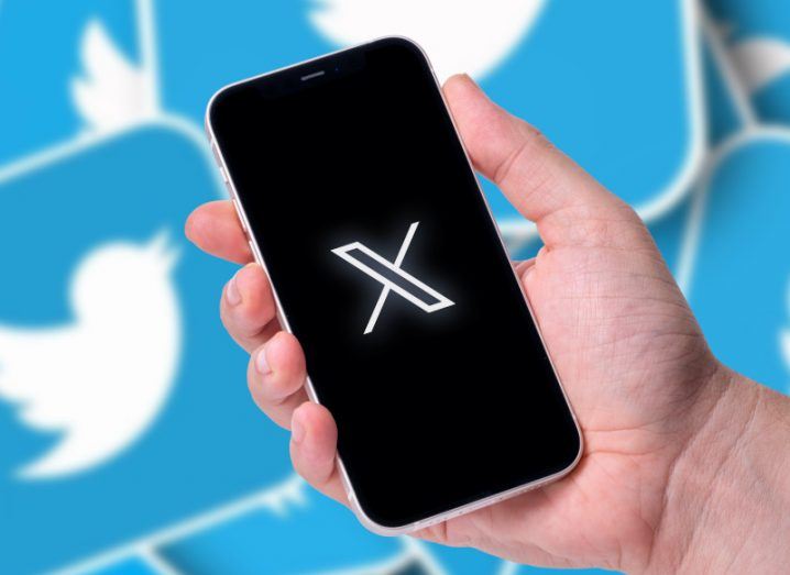 The X company logo on a smartphone screen, held in a person's hand. Multiple old Twitter logos are in the background.
