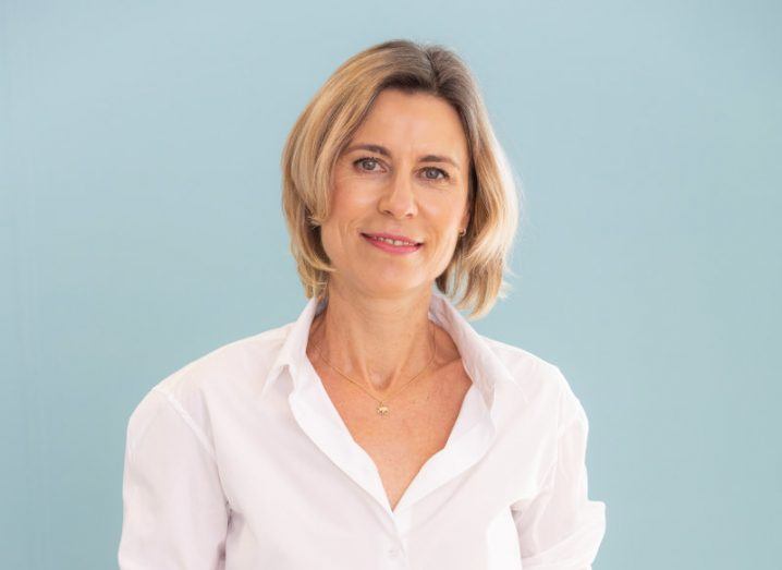 A headshot of Cathy Mauzaize. She wears a white shirt and smiles at the camera. She has short blonde hair. She stands against a pale blue background.