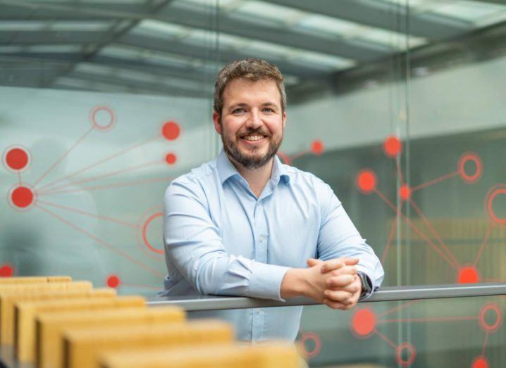 Colman O’Cathail stands leaning against a railing in front of him in a modern building with glass behind him. The glass has a red, circular design pattern on it. He is wearing a light blue shirt and his hands are crossed.