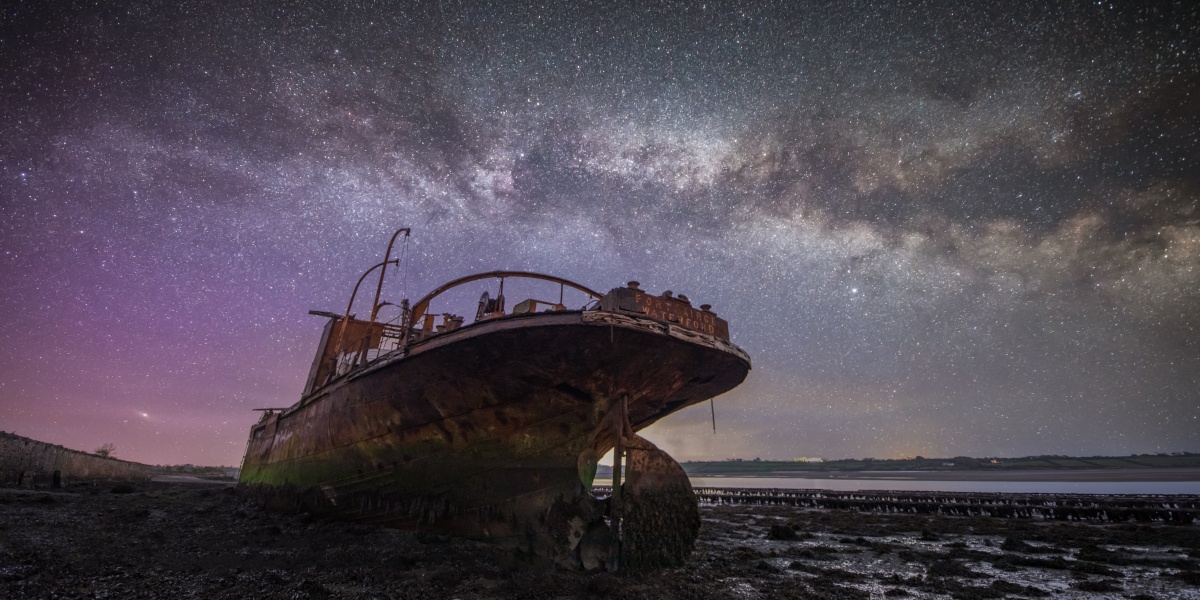 A rusty ship laying under a night sky, which shows large amounts of stars and gases from space.