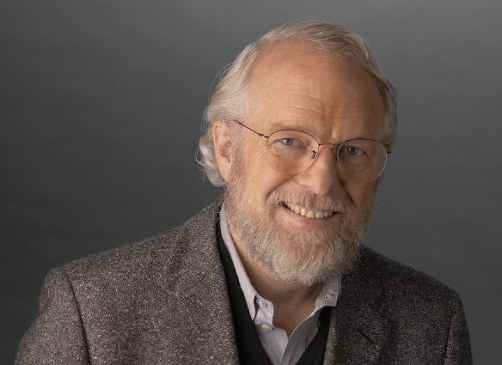 A headshot of Adobe co-founder Dr John Warnock smiling at the camera against a grey background.