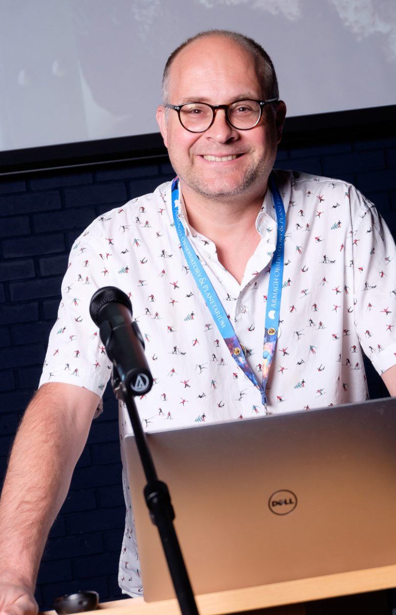 Dr Jorick Vink stand at a podium with a laptop open and a microphone in front of him. He wears a white short-sleeved shirt and a lanyard around his neck and rests his hands on either side of the podium. He smiles at the camera.