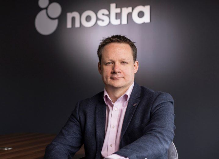 A man sitting on a chair in front of a dark wall that has the Nostra logo on it.