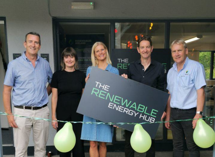 Two women and three men standing in a line in front of a ribbon with three green balloons attached to it. The woman in the centre of the image is holding a black sign that says "The Renewable Energy Hub" on it.