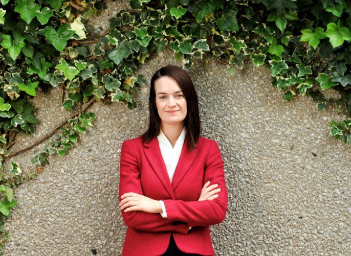 A woman wearing a red suit jacket smiles with her arms crossed in front of a stone wall with ivy growing on it.