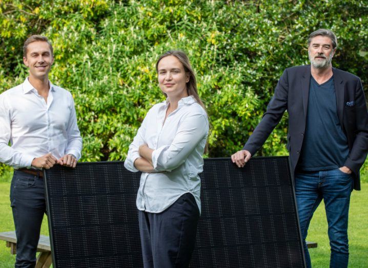 Two men and a woman stand a few metres apart in a line while propping up solar panels in the grass and smiling at the camera.