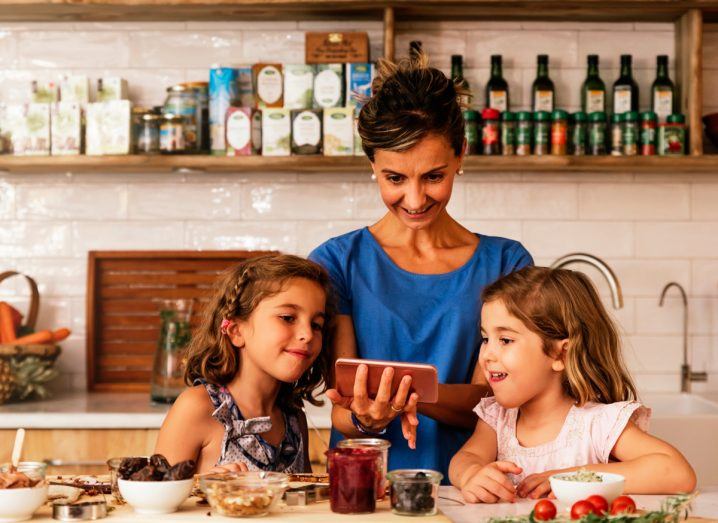 A woman with two girls staring at a phone screen in a kitchen.