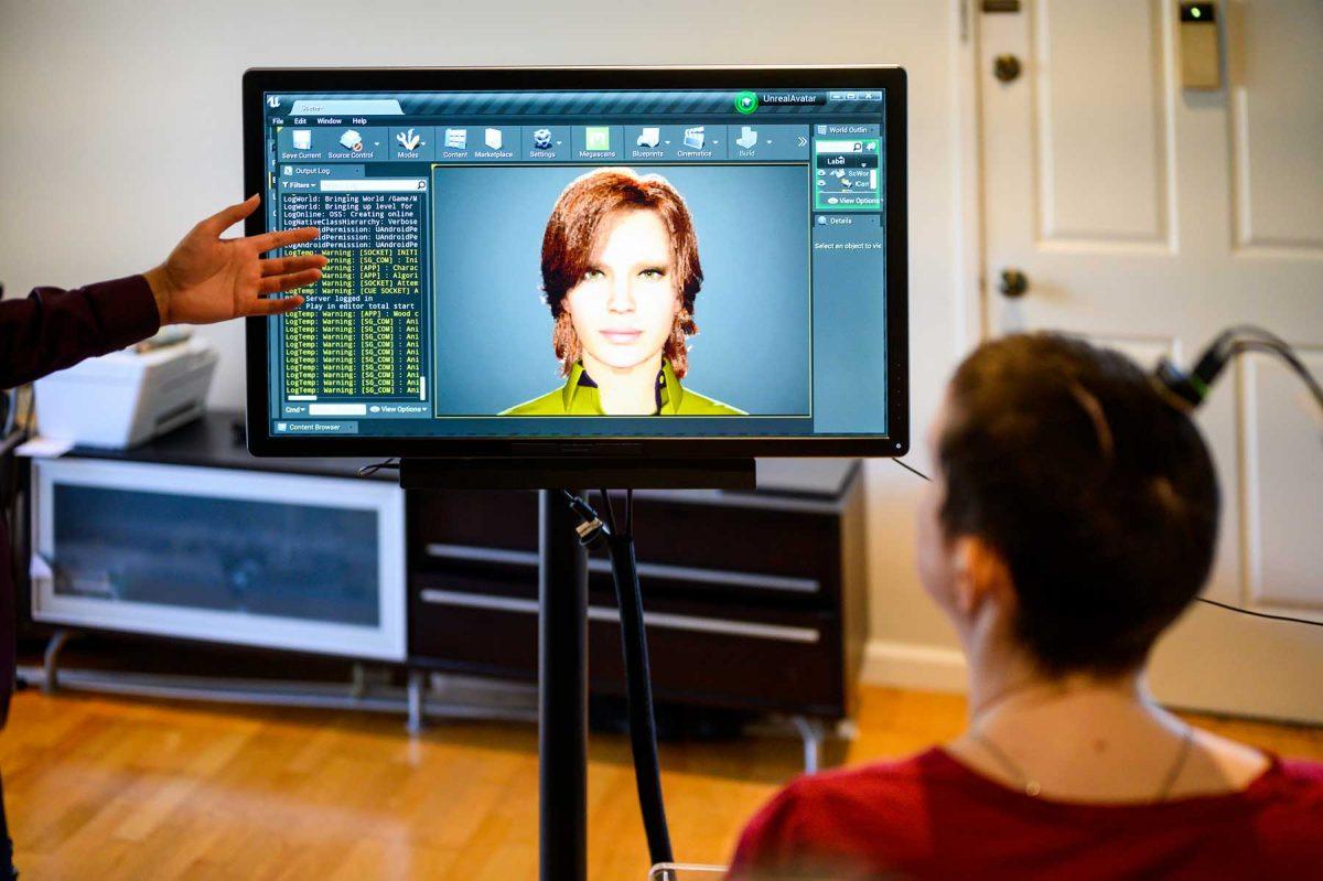 A woman sitting in front of a TV screen that has words and a digital avatar of a woman on it.