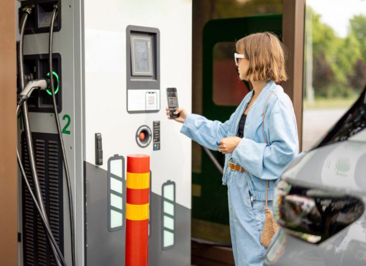 A woman stands next to an electric vehicle charging station. She is holding her phone close to the display on the charging port, trying to scan a QR code in order to pay for the service.