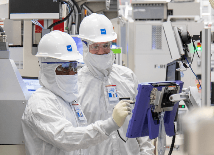 Two people in white uniforms and hardhats looking at a computer screen in a manufacturing facility. The Intel logo is on both hardhats.