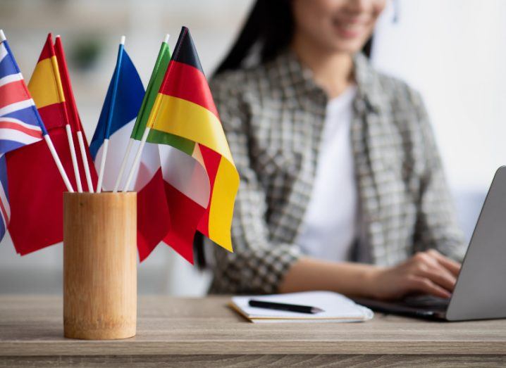 Small flags from European countries in a holder on a desk with a woman on a laptop at the desk behind.
